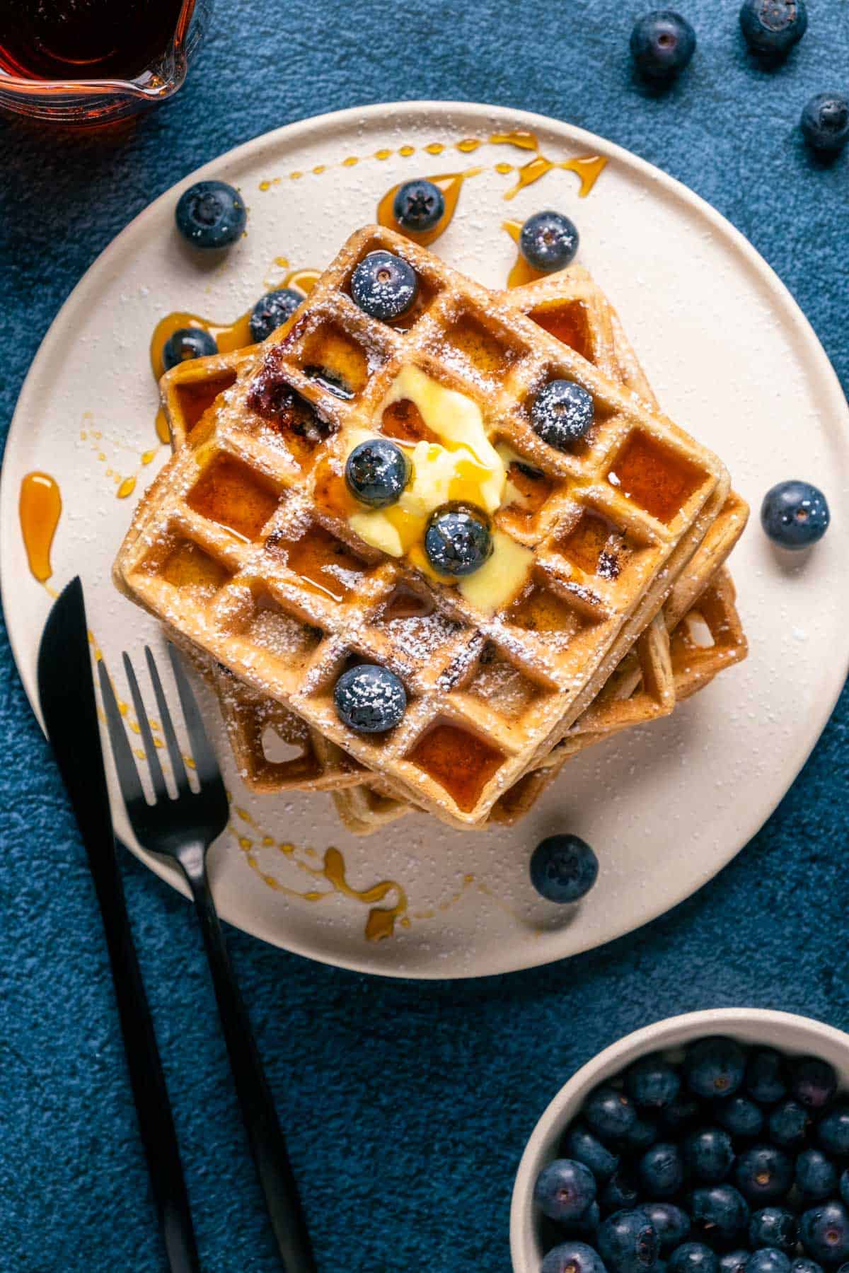 Stack of blueberry waffles topped with vegan butter, powdered sugar and syrup on a white plate.