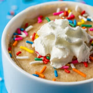 Vegan vanilla mug cake in a white mug topped with sprinkles and whipped cream.